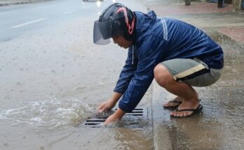 Workers clearing clogged city culvert drain using tools to remove debris, mud, and trash to restore water flow and prevent flooding.