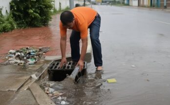 A worker removes plastic trash and debris clogging a street culvert to restore water flow and prevent flooding after heavy rain.